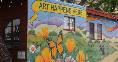 A wall painting on a side of a building features yellow with black lettering that says, "Art Happens Here." In the lower half, the painting shows an orange butterfly surrounded by orange and pink colored flowers.